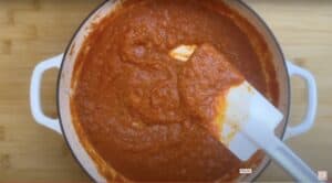 Rich homemade tomato sauce in a white pot being stirred with a spatula on a wooden surface.